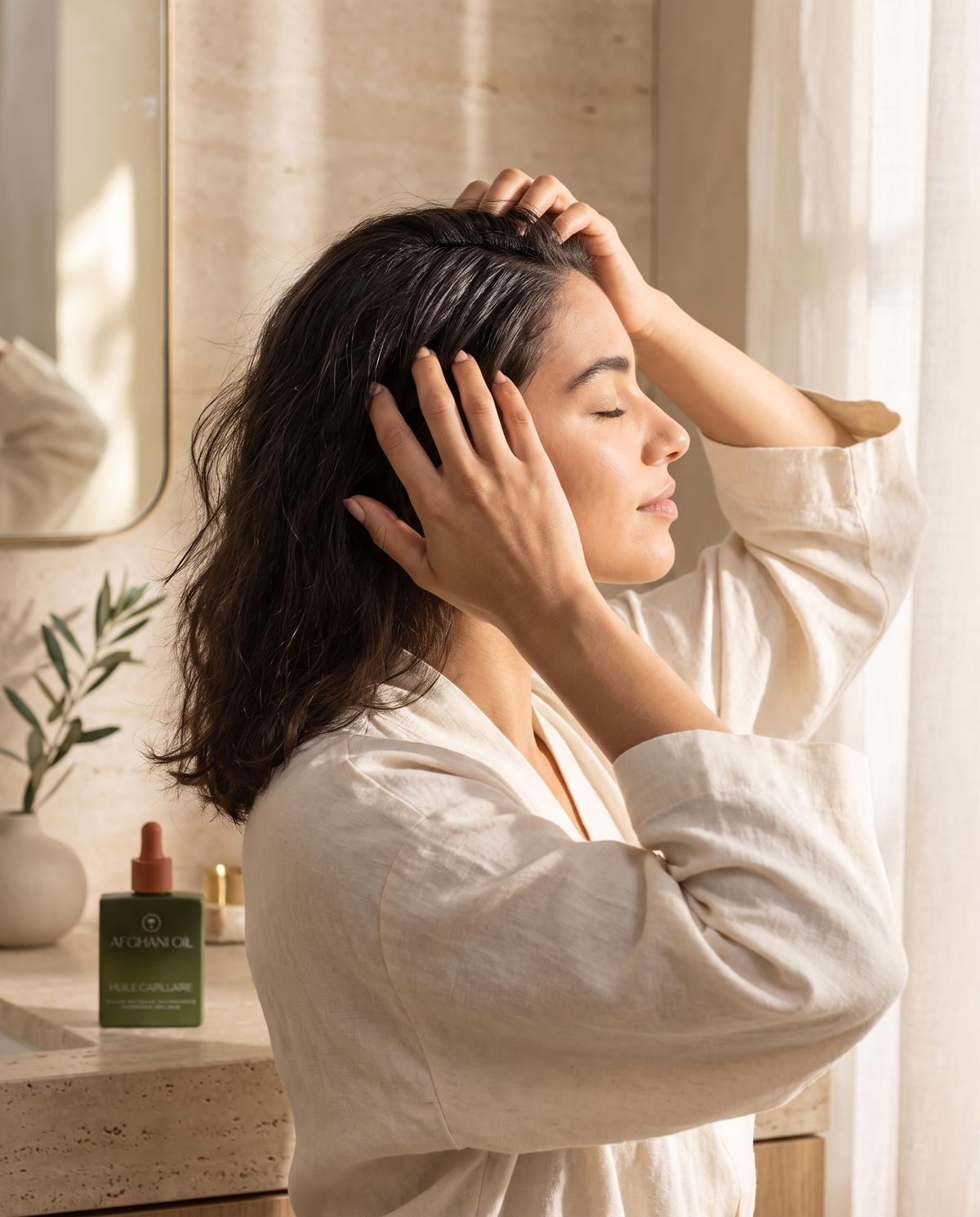 A woman in a linen robe with her fingertips in her hair, eyes closed, the Afghani Oil bottle on the counter