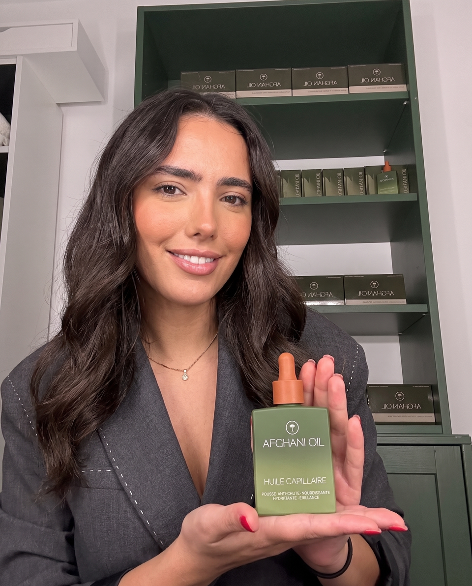 A woman in a grey blazer holding the Afghani Oil bottle in front of green shelves